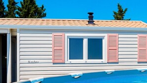 Una piscina con borde de baldosas, un edificio blanco con contraventanas rosas y una chimenea en el tejado.