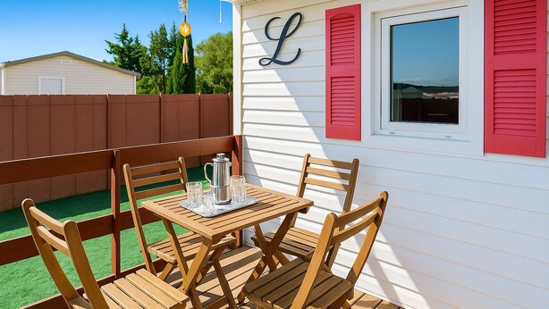 Una terraza de madera con una mesa y sillas, una jarra de metal y un buzón rojo en una casa blanca con contraventanas rojas.