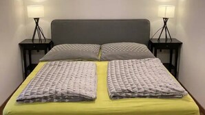 Un lit avec un matelas jaune et une literie grise, flanqué de deux tables de chevet avec des lampes, dans une pièce aux murs blancs et au plancher en bois.