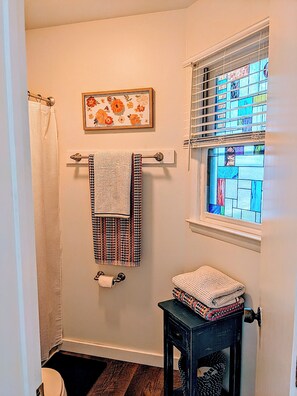 A bathroom with a shower curtain, a towel rack with towels, a framed floral artwork, a window with stained glass, and a small table with folded towels.