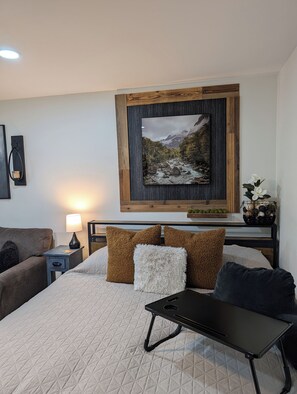 A neatly arranged bedroom with a bed, bedside table, lamp, and a framed picture on the wall. - Mountain Vibes & Morning Brews Studio retreat in Rumbling Bald-HI amenities! (Lake Lure)