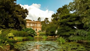 Un étang paisible, parsemé de nénuphars, entouré d’une végétation luxuriante, avec un bâtiment de style classique en arrière-plan.