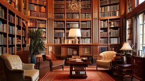 Une bibliothèque aux hautes étagères en bois remplies de livres, un lustre, des fauteuils en cuir, une moquette rouge et une grande plante en pot.