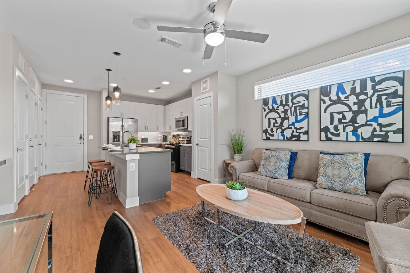 A modern kitchen with a central island, a dining area with a wooden table, and a living room with a sofa and abstract art on the wall.