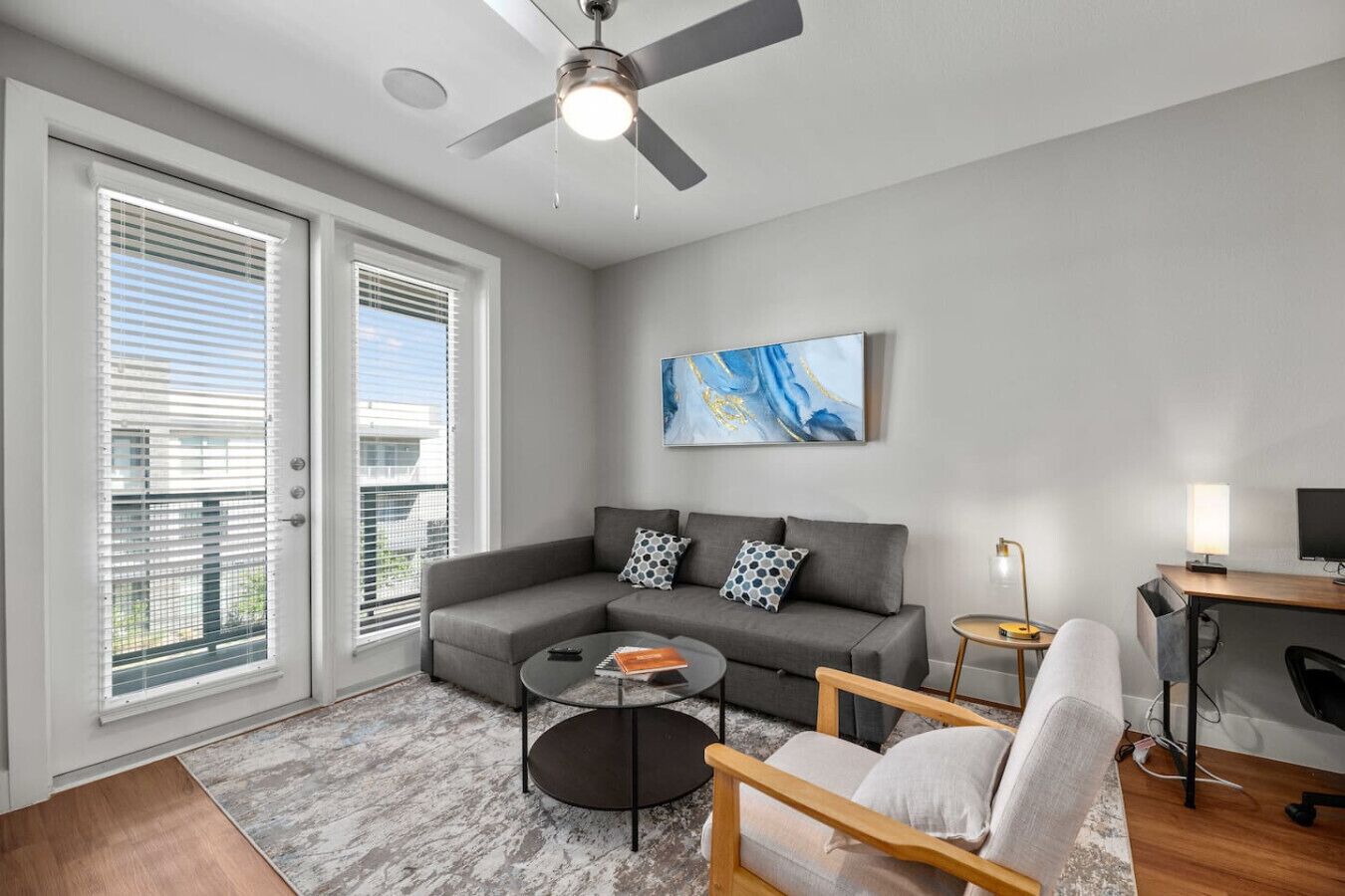 Ein modernes Wohnzimmer mit einem grauen Sofa, einem runden Couchtisch, einem Holzsesseln, einem Deckenventilator und einem Bild an der Wand.
