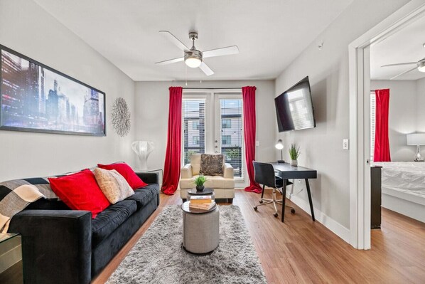 Ein modernes Wohnzimmer mit einem schwarzen Sofa, roten Kissen, einem grauen Teppich, einem weißen Sessel, einem Schreibtisch und einem an der Wand befestigten Fernseher.