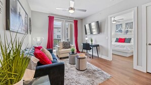 A modern living room with a sofa, coffee table, and a TV. There is a bed with red bedding and a door leading to another room. - Stylish Luxury King Bed | Gym+Pool+Stockyards (Fort Worth)
