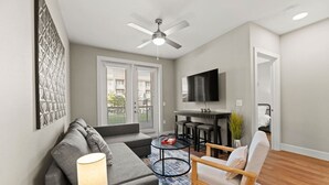 A modern living room with a grey sofa, a round coffee table, a wooden chair, a TV, and a ceiling fan. - Sleeps 10 w/King Bed+Gym+Free Parking+Stockyards (Fort Worth)