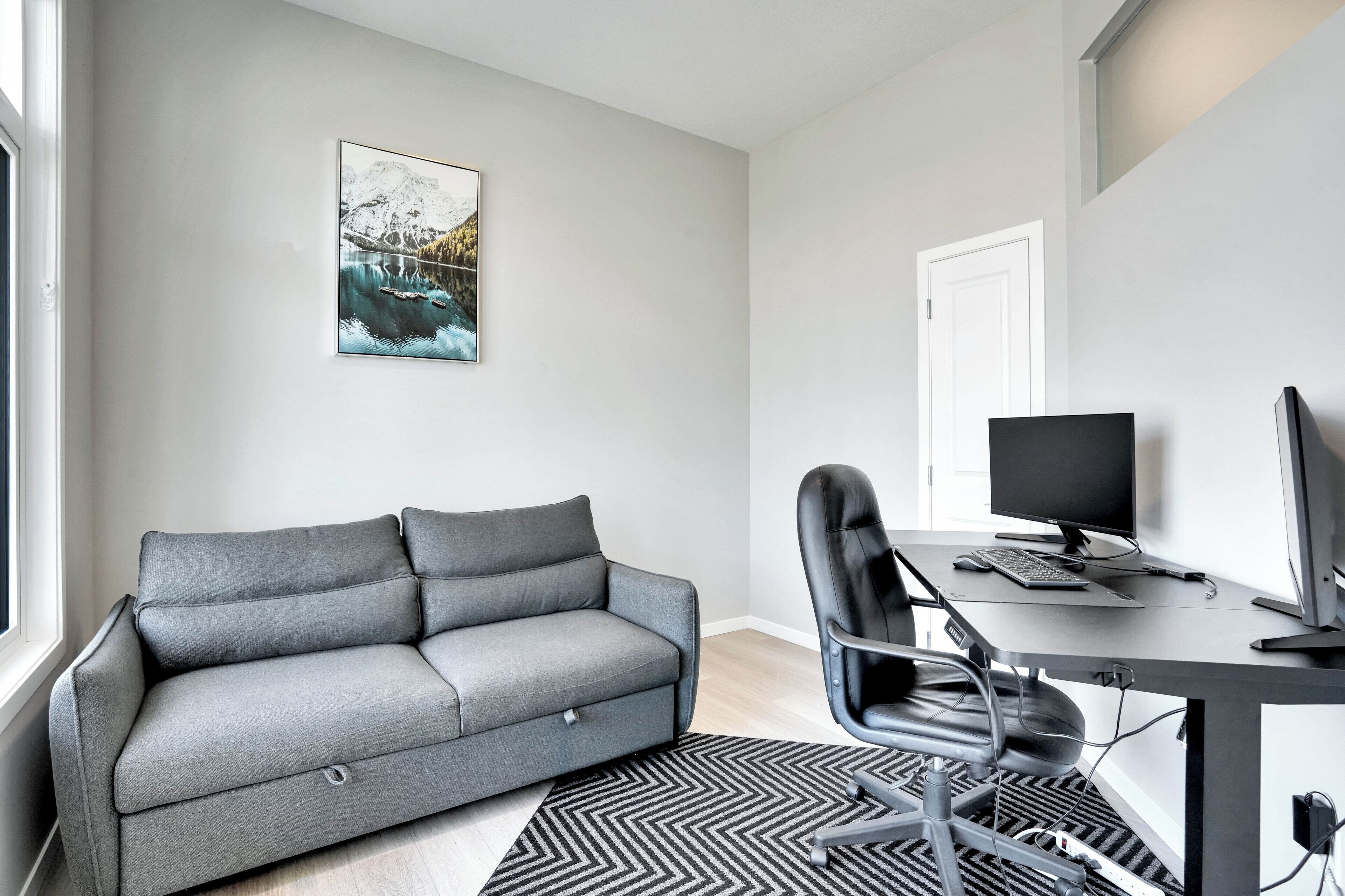 Ein modernes Wohnzimmer mit einem grauen Sofa, einem schwarzen Bürostuhl, einem Schreibtisch mit Computer und einem schwarz-weißen Teppich.