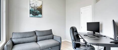 Ein modernes Wohnzimmer mit einem grauen Sofa, einem schwarzen Bürostuhl, einem Schreibtisch mit Computer und einem schwarz-weißen Teppich.