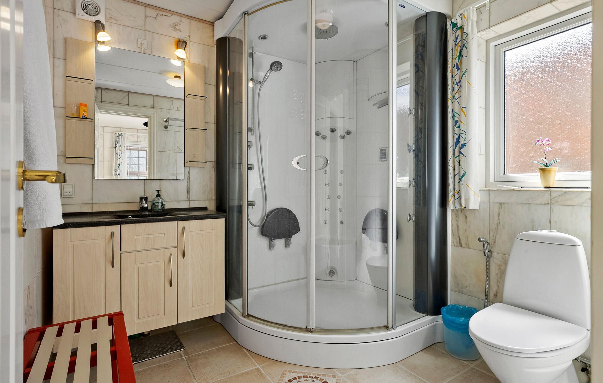 Een moderne badkamer met een douchecabine van glas, een toilet, een wastafel met spiegel, en een raam met een plant in pot.