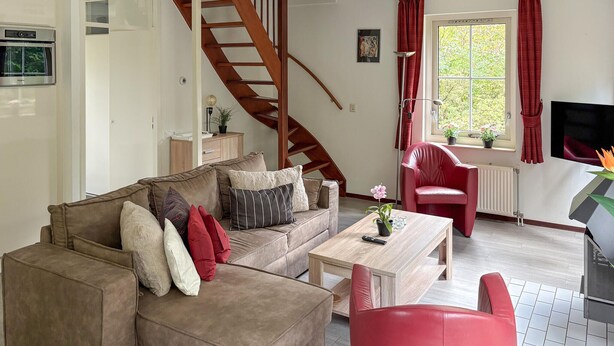 Een woonkamer met een bruine bank, een rode fauteuil, een houten salontafel en een trap.