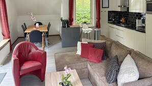 Een moderne woonkamer met een rode leren fauteuil, een bruine bank en een houten salontafel. Er is een eethoek met een tafel en stoelen, en een keuken met een zwart betegelde achterwand en witte kasten.