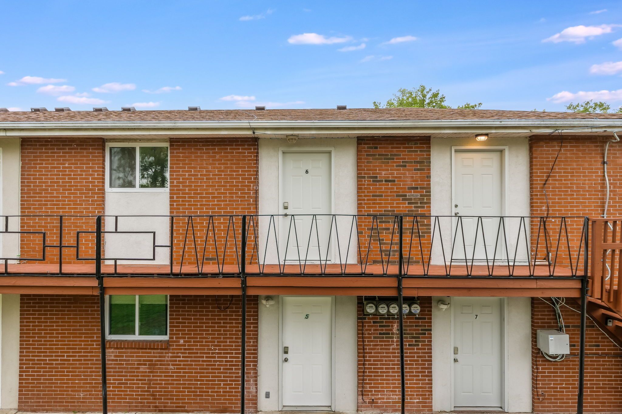 A two-story brick building with numbered doors, a balcony with a black railing, and a clear blue sky.