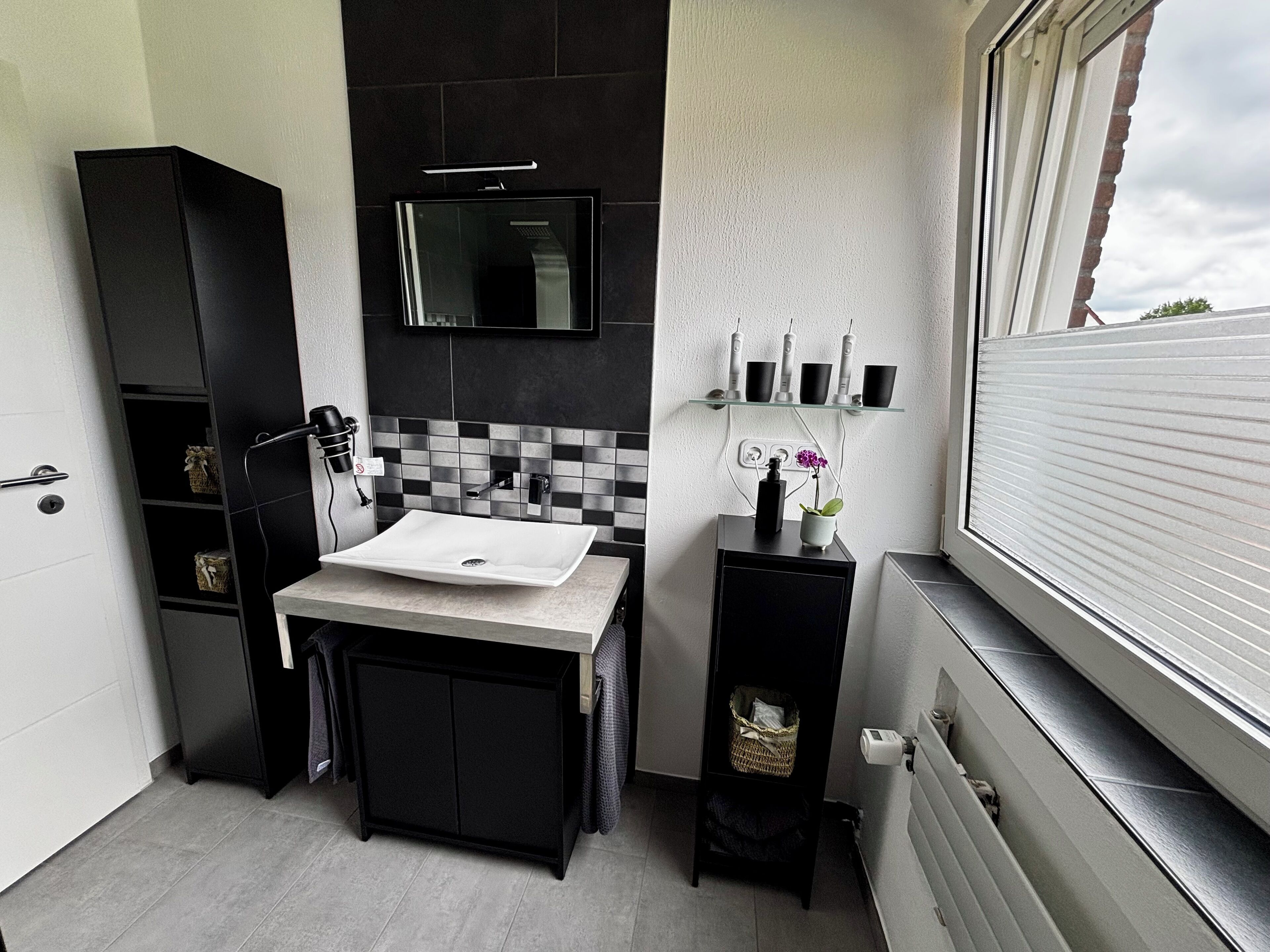 Ein modernes Badezimmer mit einem schwarzen Schrank, einem weißen Waschbecken und einem Spiegel darüber. Eine Fenster mit Jalousien befindet sich auf der rechten Seite.