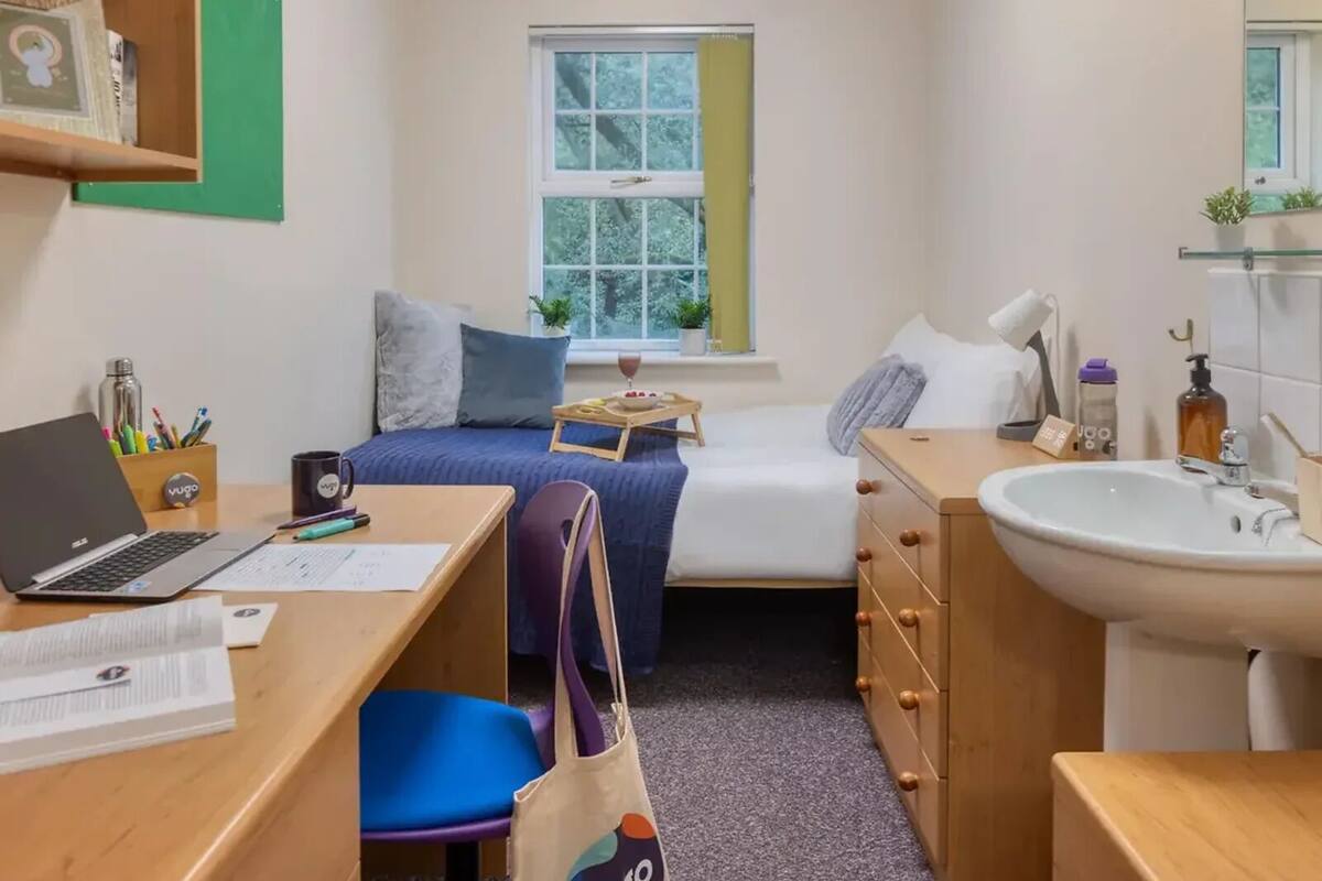 Une petite chambre de dortoir comprenant un lit, un bureau, une chaise et un petit lavabo.