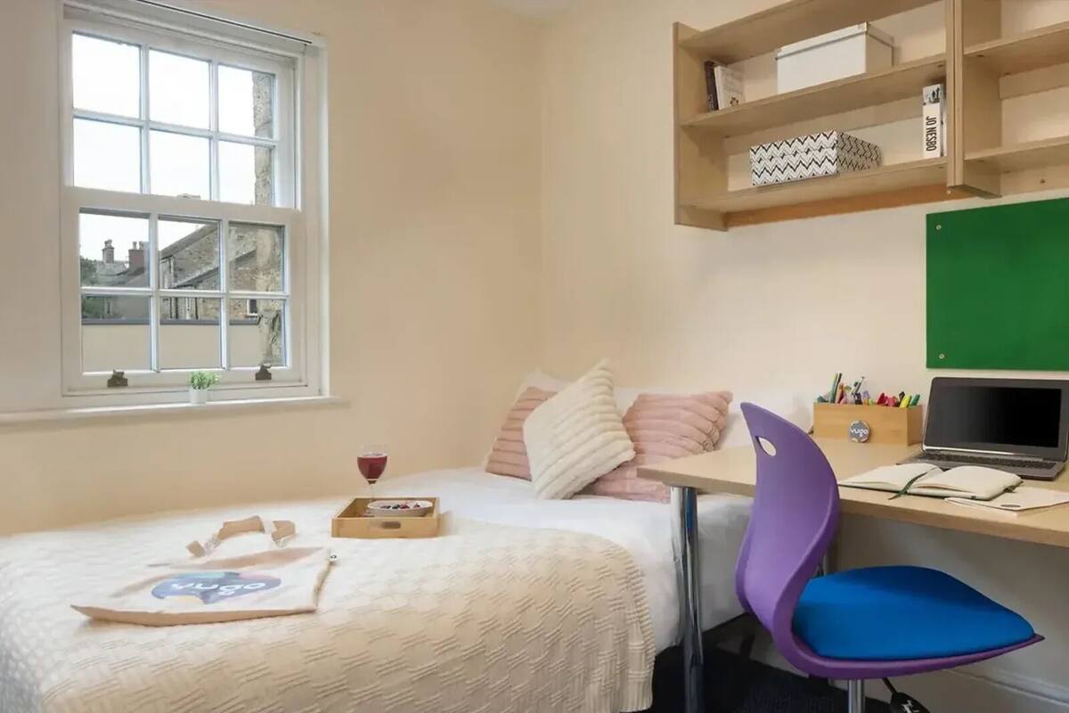 Une chambre à coucher avec un lit, un bureau avec un ordinateur portable, une chaise et une fenêtre donnant sur des immeubles.