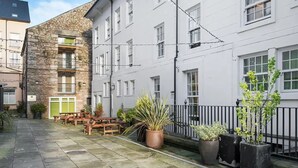 A courtyard with stone flooring, wooden benches, potted plants, and a white building with multiple windows and balconies. - Student Only Cable Street (Lancaster)