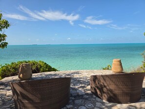 A seaside view with two wicker chairs and a terracotta pot on a stone patio.