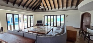 A spacious living area with a sofa, a coffee table, and a TV. There is a large window providing a view of the sea.