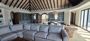 Amplia sala de estar con un sofá seccional gris grande, una mesa de comedor con sillas y una cocina con armarios azules y un ventanal.