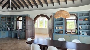 Ein geräumiges Zimmer mit hoher Decke, einer Essecke mit weißen Stühlen, einer Couch mit Kissen und eingebauten Regalen mit diversen Gegenständen.