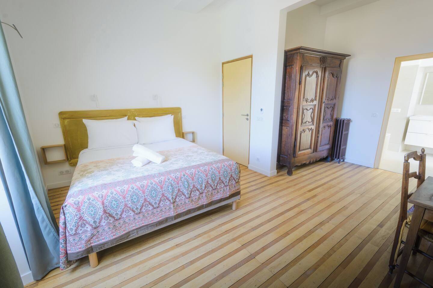 A bedroom with a wooden floor, a bed with a patterned bedspread, a wooden wardrobe, and a chair.