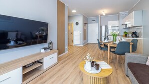Ein modernes Wohnzimmer mit einem Flachbildfernseher, einem Holztisch mit Dekoration, einer Essecke mit Holztisch und -stühlen sowie einer Küche mit weißen Schränken und einem Herd.