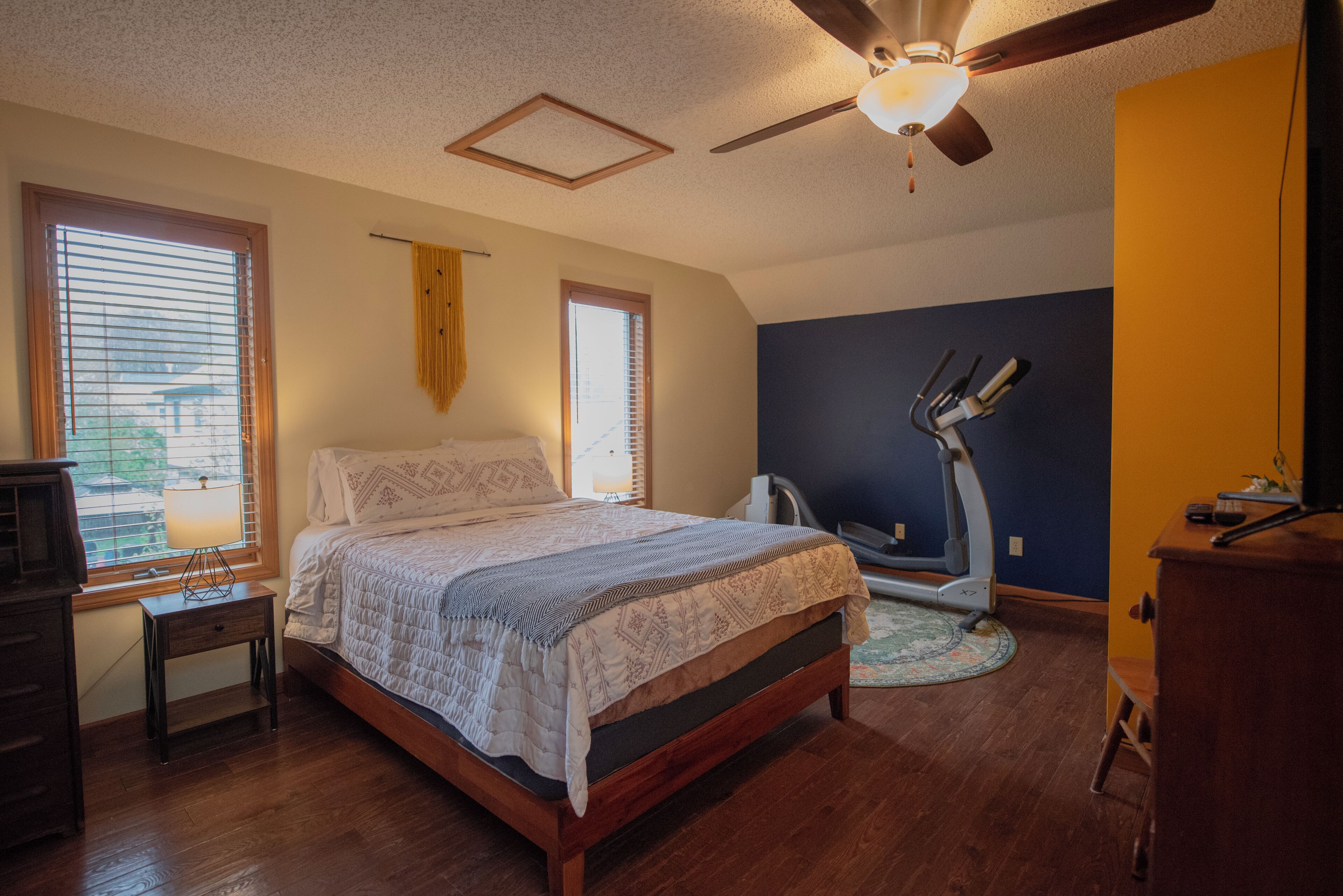 A bedroom with a bed, a ceiling fan, a treadmill, and a window with blinds.