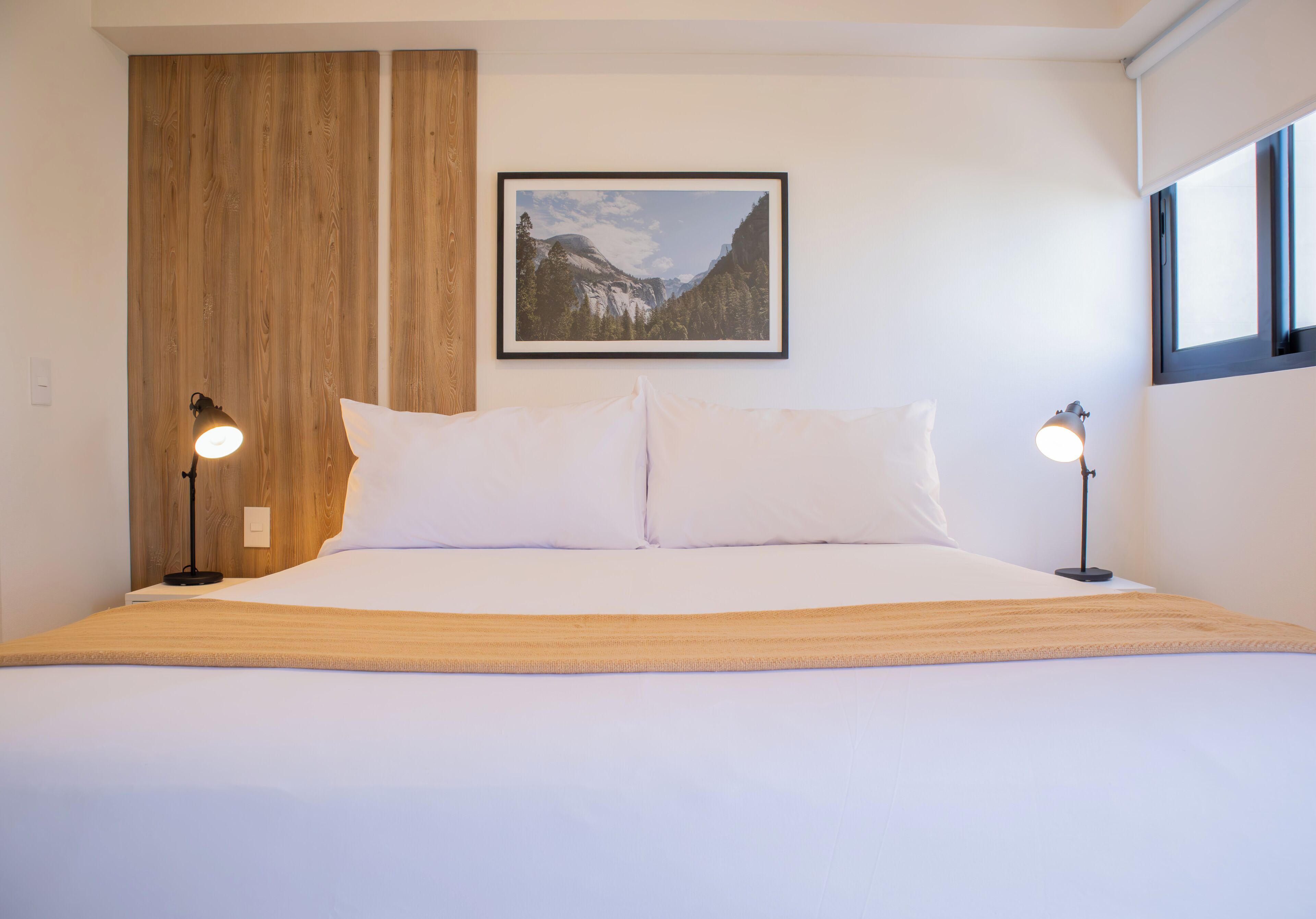 Una cama bien hecha con sábanas blancas y dos lámparas de noche. Un cuadro enmarcado de un paisaje montañoso cuelga sobre la cama.