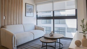 Una habitación de hotel moderna con un sofá blanco, una mesa redonda y un ventanal con persianas.