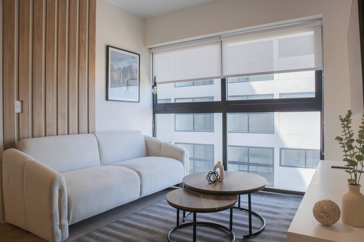 A modern hotel room with a white sofa, a round table, and a large window with blinds.