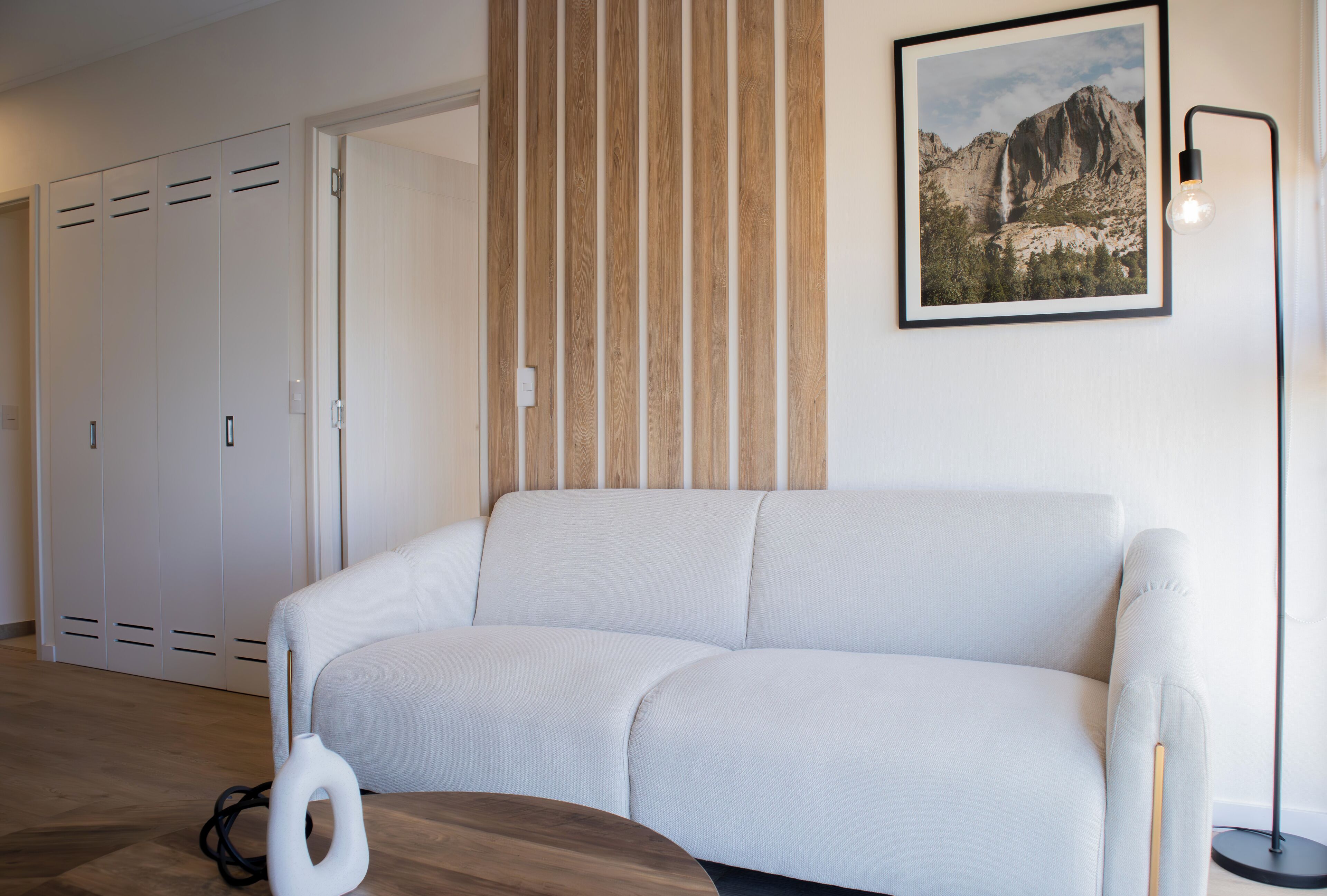 A modern living room with a white sofa, wooden slat wall paneling, a framed picture of a mountain, and a floor lamp.