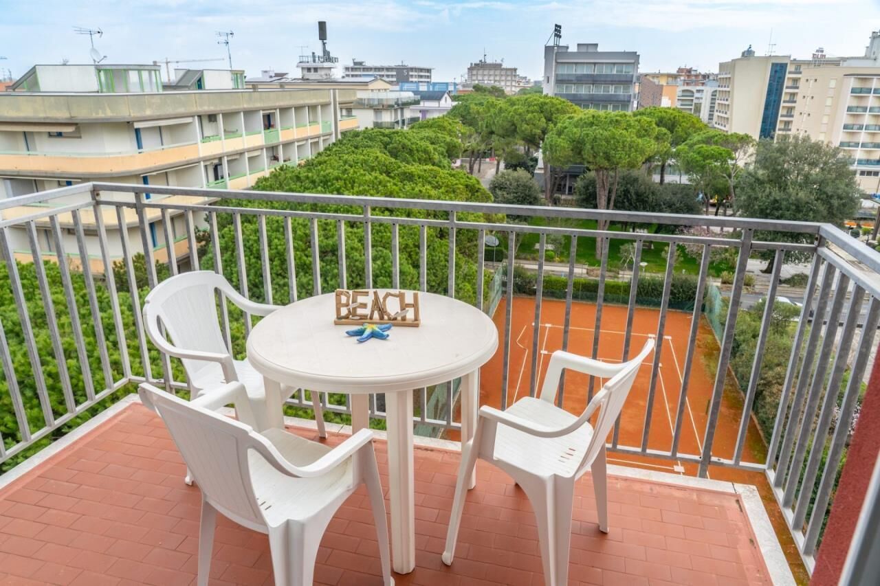 Un balcón con una mesa y sillas, un cartel que dice "BEACH" y vista a una cancha de tenis y edificios circundantes.