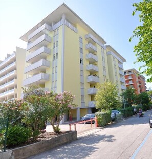 A multi-story residential building with balconies, a parking area, and surrounding greenery.