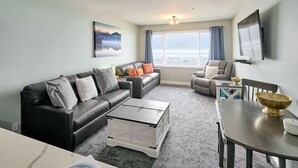 A modern living room with a sofa, coffee table, and dining table. There is a large window with curtains, a flat-screen TV, and a painting on the wall. - Remodeled Oceanfront Suite - Indoor Pool and Ocean Views! (Lincoln City)
