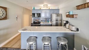 A modern kitchen with grey cabinets, a central island with bar stools, stainless steel appliances, and a decorative clock on the wall. - Remodeled Oceanfront Suite - Indoor Pool and Ocean Views! (Lincoln City)