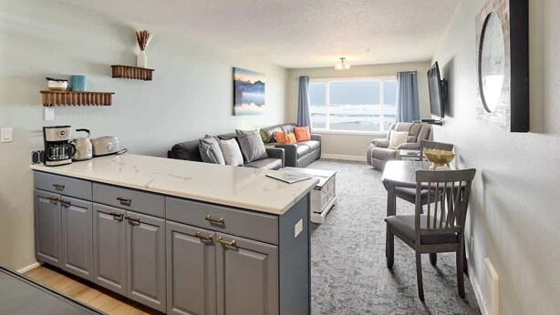 A modern kitchen with grey cabinets, a central island, and a dining area with a table and chairs. - Remodeled Oceanfront Suite - Indoor Pool and Ocean Views! (Lincoln City)