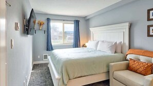 A bedroom with a bed, a chair, a TV mounted on the wall, and a window with curtains. - Remodeled Oceanfront Suite - Indoor Pool and Ocean Views! (Lincoln City)