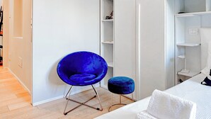 Une chambre à coucher moderne avec un lit, une chaise bleue, un petit pouf et une armoire blanche.