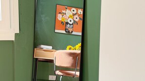A mirror reflecting a room with a green wall, a wooden desk, a chair, and a painting of flowers.