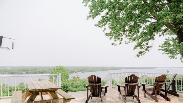 A wooden deck with a picnic table and Adirondack chairs overlooking a body of water. - Bayview House at Lake on the Mountain (Prince Edward)