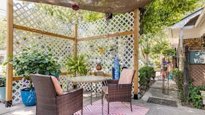 Un patio cubierto con mobiliario de mimbre y una pequeña mesa, rodeado por un cerramiento ajardinado y plantas en macetas.