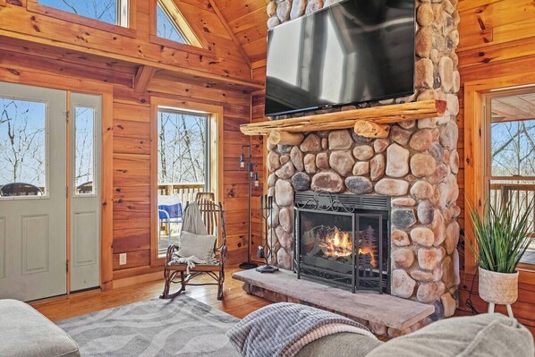 A cozy cabin interior with a stone fireplace, a television mounted above it, and a rocking chair by the window.