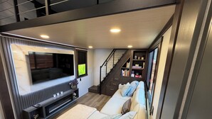 A modern living room with a large flat-screen TV, a sofa with cushions, a coffee table, and a staircase leading to a bookshelf.