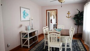 Un comedor con una mesa de madera, sillas blancas, un tapete azul y un ventilador de techo.