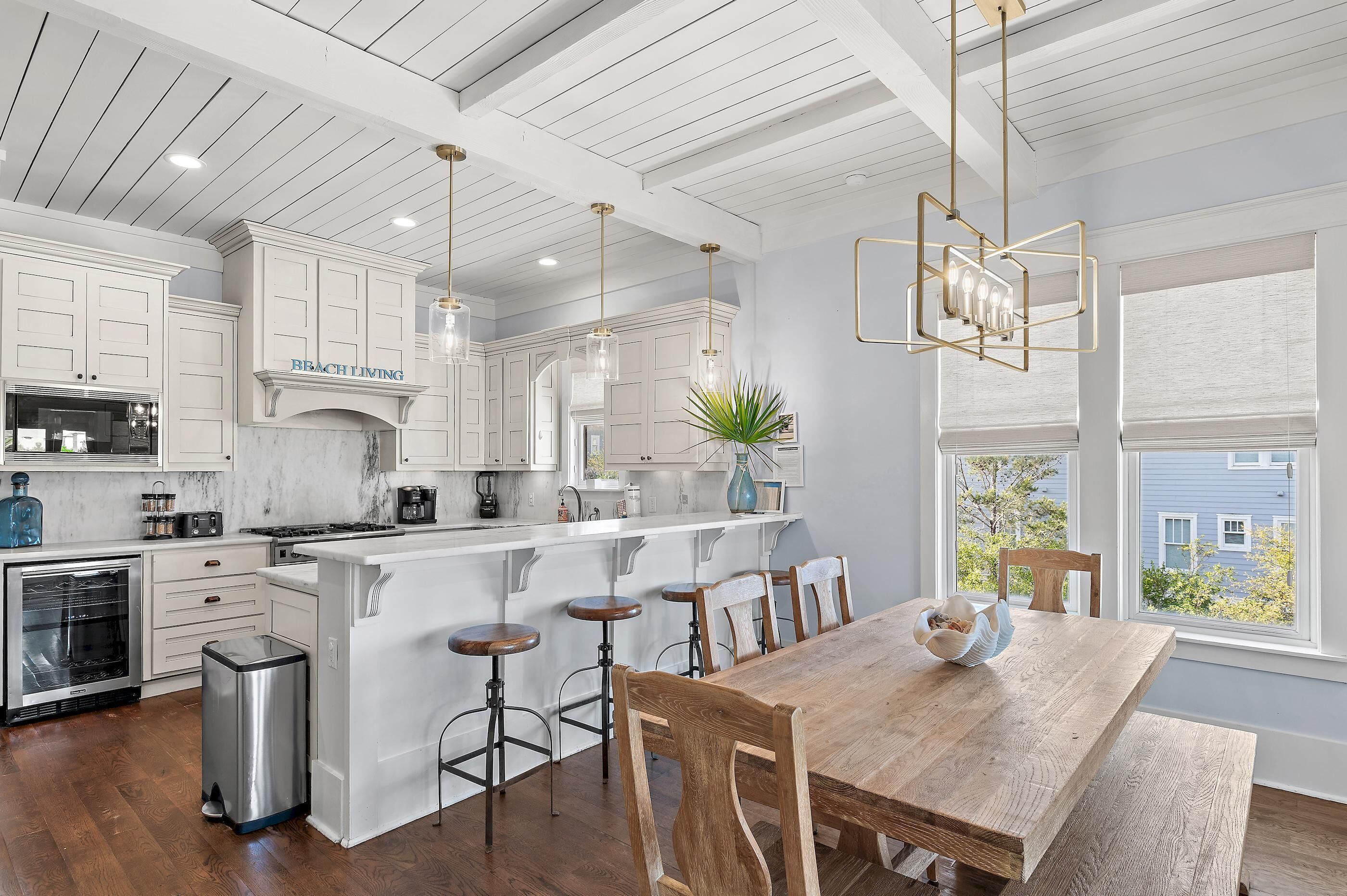 A kitchen with white cabinetry, a wooden dining table, bar stools, and a view through large windows.