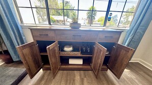 Un meuble en bois aux portes ouvertes, révélant un espace de rangement, une fenêtre donnant sur des arbres et des bâtiments, et un rideau bleu.