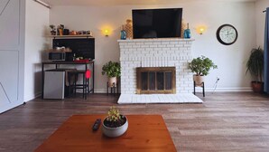 Un salon doté d’une cheminée en briques blanches, d’un téléviseur à écran plat fixé au-dessus, d’un coin repas avec une table et des chaises, et d’une horloge au mur.
