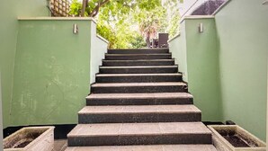 Eine Treppe führt zur Hauseingang eines Gebäudes mit grünen Wänden, im Hintergrund ist ein Baum zu sehen.